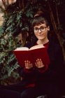Молода усміхнена елегантна жінка в окулярах з книгою, що сидить на лавці в міському саду і дивиться на камеру — стокове фото