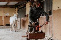 Дорослий фермер використовує молоток і щипці для кування гарячої підкови на портативній ковадлі поблизу стайні на ранчо — стокове фото