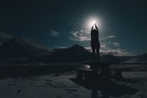 Silhouette of woman standing on table on river shore with upped hands to moon in sky at night — Stock Photo