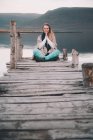Young woman doing meditation while sitting on dock near amazing water surface between hills — Stock Photo