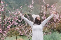 Mulher excitada em suéter e chapéu levantando os braços enquanto está em pé no incrível campo de primavera e desfrutando de liberdade — Fotografia de Stock