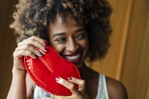 Smiling black female with curly hair looking away while holding bright lip shaped case at home — Stock Photo