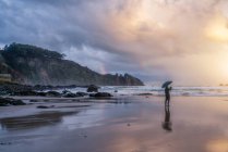 Задній вид людини з парасолькою стоячи на березі моря навколо валунів і хлюпалися хвиль на sundown — стокове фото