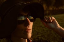 Junge sinnliche Brünette trägt schwarzen Hut und Accessoires, während sie im Sonnenlicht mit Sonnenbrille posiert — Stockfoto