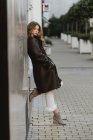 Stylish young woman in vintage leather coat leaning on wall on city street — Stock Photo