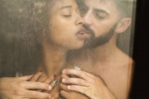 Schöner hispanischer Kerl, der die verführerische afrikanisch-amerikanische Frau anfasst und küsst, während er zu Hause hinter dem nassen Fenster steht — Stockfoto