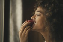 Seductive African American female with curly hair eating ripe strawberry at home — Stock Photo