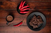 Mug of fresh hot drink placed on lumber tabletop near plate with pieces of chocolate and chili pepper — Stock Photo