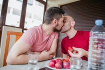 Affectionate gay couple eating strawberries at table in kitchen — Stock Photo