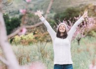 Emocionada mujer en suéter y sombrero levantando brazos mientras está de pie en el increíble campo de primavera y disfrutando de la libertad - foto de stock