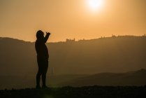 Silueta de la persona de pie en el campo en la espalda brillante iluminado por el cielo puesta del sol, Italia - foto de stock
