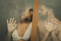 Handsome Hispanic guy touching and kissing seductive African American woman in lace bra while standing behind wet window at home — Stock Photo