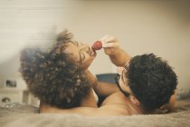 Bearded guy feeding cheerful girlfriend with fresh strawberry while lying on comfortable bed together — Stock Photo