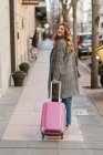 Приваблива жінка в модному пальто потягнувши рожевий чемодан і посміхаючись під час ходьби по міській вулиці і дивлячись на камеру — стокове фото