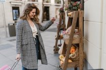 Jeune femme élégante souriante avec valise regardant les épiceries sur des étagères en bois près du magasin dans la rue de la ville — Photo de stock