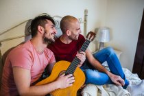 Cheerful gay couple playing guitar in bedroom — Stock Photo