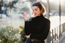 Joven dama en traje de moda exhalando humo mientras fuma cigarrillo en día soleado en la calle de la ciudad - foto de stock
