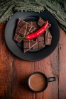 Mug of fresh hot drink placed on lumber tabletop near plate with pieces of chocolate and chili pepper — Stock Photo