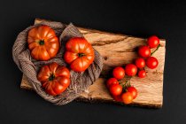 Fresh ripe tomatoes and fabric napkin on piece of wood on black background — Stock Photo
