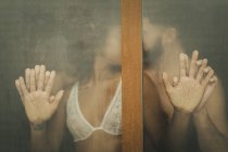 Guapo chico hispano tocando y besando seductora mujer afroamericana en sujetador de encaje mientras está de pie detrás de una ventana húmeda en casa - foto de stock