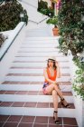 Joven mujer china con clase en sombrero de panama y gafas de sol sentado en las escaleras y disfrutando de un día de verano brillante - foto de stock