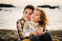Веселые и симпатичные мальчик и девочка улыбаются и обнимают друг друга на пляже — стоковое фото