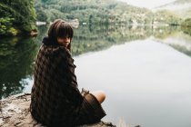 Woman sitting with blanket near lake and mountains — Stock Photo