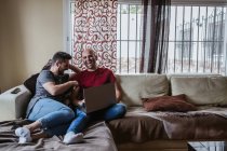 Весела гей пара використовує ноутбук, розслабляючись на дивані — стокове фото