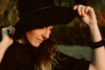 Young sensual brunette wearing black hat and accessories while posing in sunlight keeping eyes closed — Stock Photo