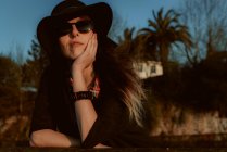 Пряма жінка в модних сонцезахисних окулярах з чорним капелюхом, що спирається на руку на сонячне світло — стокове фото