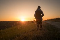 Visão traseira da pessoa na jaqueta com as mãos em bolsos de pé em campos verdes da Itália ao pôr do sol — Fotografia de Stock