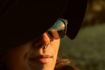 Giovane bruna sensuale con cappello nero e accessori mentre posa alla luce del sole con occhiali da sole — Foto stock