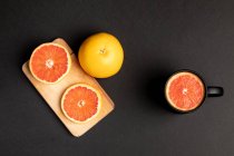 Sliced and whole grapefruits on black background near wooden board and mug — Stock Photo