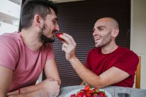 Cheerful gay couple eating strawberries at table in kitchen — Stock Photo