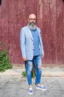 Vue de face d'un hipster barbu mature à la mode debout contre une porte rouge vintage à l'extérieur tout en regardant la caméra — Photo de stock