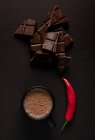 Des morceaux de chocolat délicieux placés près de tasse noire de boisson chaude avec du piment sur fond sombre — Photo de stock