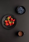 Various fresh berries and mug of aromatic hot beverage for breakfast on black background — Stock Photo