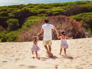Обратный вид анонимного отца с двумя дочерьми, гуляющими по песчаной дюне в яркий летний день — стоковое фото