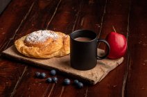 Manzana roja fresca y arándanos maduros colocados en la mesa de madera cerca de taza de bebida caliente aromática y delicioso bollo para el desayuno por la mañana - foto de stock