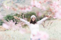 Захоплена жінка в светрі і капелюсі піднімає руки, стоячи в дивовижній весняній сільській місцевості і насолоджуючись свободою — стокове фото