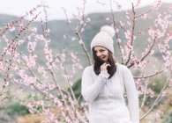 Donna sorridente in maglione e cappello in piedi vicino all'albero in fiore nella campagna primaverile — Foto stock
