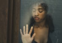 Pretty African American woman with closed eyes touching wet glass surface while standing behind window — Stock Photo