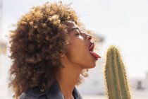 Attractive African American female with closed eyes pretending to lick prickly cactus against blurred background of street on sunny day — Stock Photo