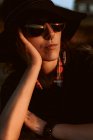 Пряма жінка в модних сонцезахисних окулярах з чорним капелюхом, що спирається на руку на сонячне світло — стокове фото