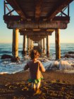 Kind von hinten unter einem Pier am Strand bei Sonnenuntergang — Stockfoto