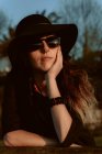 Pensive woman wearing trendy sunglasses with black hat leaning on hand in sunlight — Stock Photo