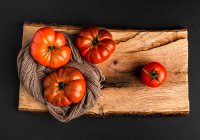 Pomodori freschi maturi e tovagliolo di tessuto su pezzo di legno su sfondo nero — Foto stock