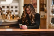 Красивая женщина в модном наряде смеется и просматривает современный смартфон, сидя за столом в стильном кафе — стоковое фото