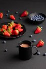 Различные свежие ягоды и кружка ароматного горячего напитка на завтрак на черном фоне — стоковое фото
