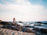 Анонимная маленькая девочка сзади стоит на пляже на песке, глядя на море — стоковое фото